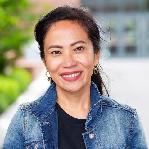 Woman with olive-colored skin, long dark hair and big, bright smile. She is wearing red lipstick, black shirt and denim shirt with popped collar.